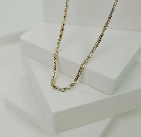 Yellow gold wheat or spiga braided chain draped over white display blocks, clasp hanging at the bottom of the loop — stand-in photograph